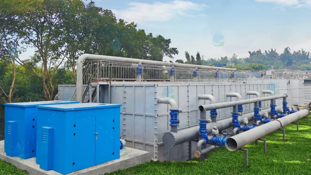 A large, multi-stage grey water storage and treatment tank on a grassy area, integrated with a network of grey and blue pipes and a blue equipment cabinet, possibly connected to a high-capacity 8 inch borehole pump extraction point.