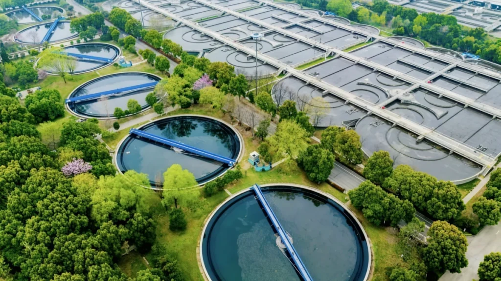 Aerial view of a massive water treatment plant featuring numerous circular clarification tanks and rectangular aeration basins, surrounded by trees, capable of processing groundwater extracted by an 8 inch submersible well pump network.