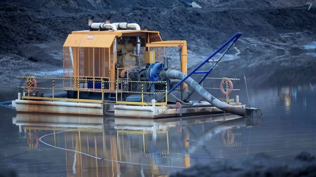 A floating, heavy-duty industrial pump barge with an enclosed yellow engine compartment and a large blue pump, actively dewatering and extracting sediment from a water source, suitable for applications alongside an 8 inch submersible well pump.