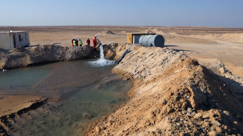 A team of workers in safety gear and hard hats supervising a powerful, constant water discharge from a pipe into a large desert reservoir, drawing water from a borehole with an industrial-grade submersible motor.