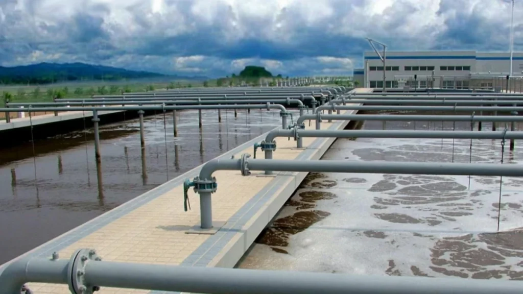 An extensive array of foamy, actively mixed wastewater aeration tanks at a treatment plant, featuring a complex network of overhead grey pipelines and control valves connected to large submersible motors.
