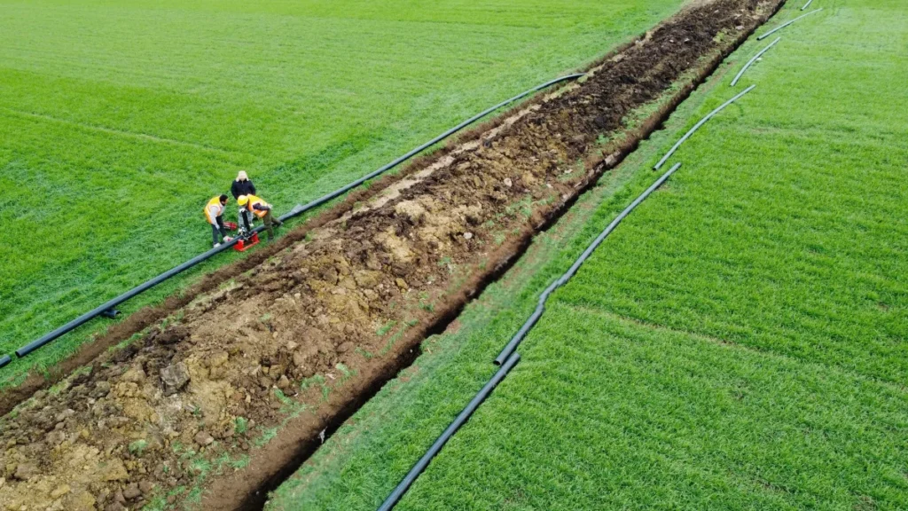 An aerial view showing two workers installing segments of pipeline into a long trench spanning a large, vibrant green agricultural field, preparing the irrigation infrastructure to be powered by a submersible motor.