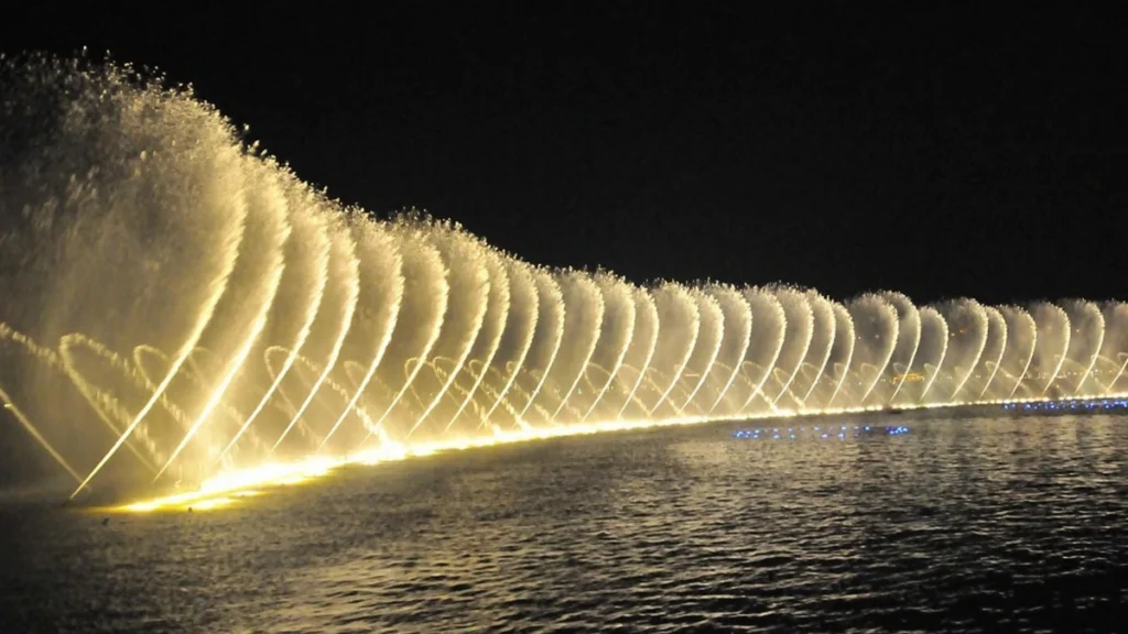 A spectacular nighttime view of a large-scale illuminated musical fountain system, with many synchronized water jets creating golden wave patterns across a lake, forced by multiple heavy-duty submersible electric motors.